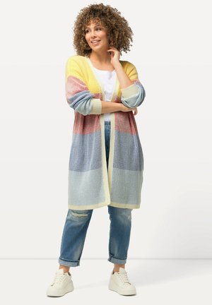 Cardigan lungo a maglia aperta in giallo, rosso, blu e crema. Caratterizzato da una vestibilità rilassata, spalle scese e polsini a coste. Indossato sopra un top bianco.