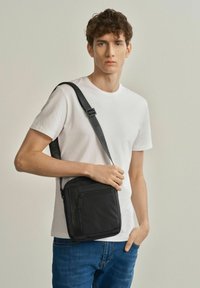 Czarna torba na ramię o gładkiej fakturze, z zamykaną kieszenią i regulowanym paskiem, noszona przez osobę w prostym białym T-shircie i niebieskich dżinsach.