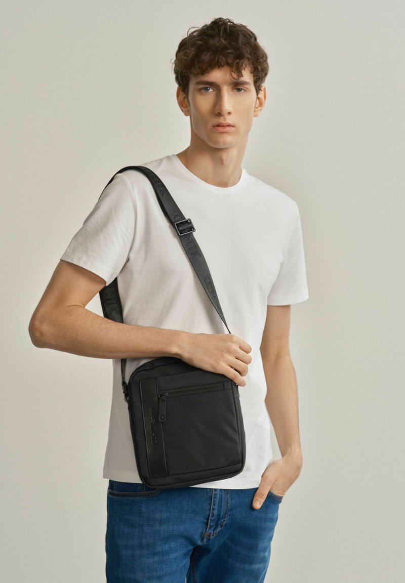 Czarna torba na ramię o gładkiej fakturze, z zamykaną kieszenią i regulowanym paskiem, noszona przez osobę w prostym białym T-shircie i niebieskich dżinsach.