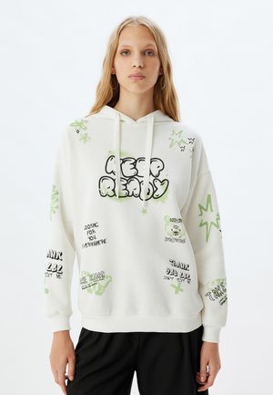 Sweat à capuche blanc avec des impressions graphiques vertes et noires, y compris du texte et des accents de dessin animé. Comprend des manches longues, une poche frontale et une capuche à cordon.