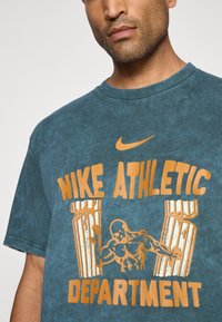 Nike atletisk T-shirt i mørkeblå med orange grafisk tryk. Har rund halsudskæring, korte ærmer og et tekstureret stof med et vintage look.