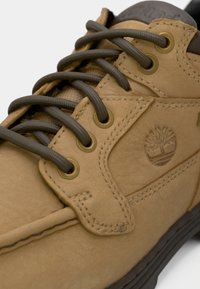 Gros plan d'une chaussure en cuir fauve avec des lacets marron foncé et un logo d'arbre en relief sur le côté près du col noir rembourré.