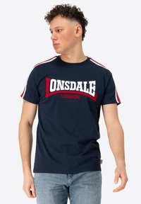 Námořnická modrá bavlněná tričko s výrazným bílým a červeným logem "Lonsdale London", krátké rukávy s červenými a bílými pruhy na ramenou.