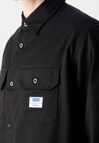 Chemise noire en tissu lisse avec un col pointu, deux poches poitrine et une étiquette bleue portant "HUGO." Boutons noirs.