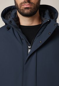 Mężczyzna ubrany w granatową kurtkę z kapturem, zapinaną na zamek i guziki, mający na sobie czarną bluzkę, z zarostem na twarzy i zamkniętymi ustami.