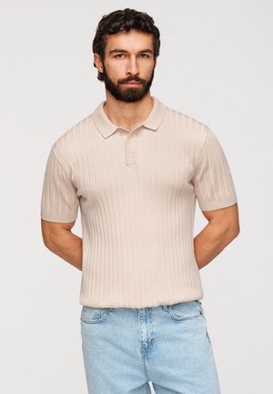 Homme barbu aux cheveux bouclés foncés, portant un polo beige côtelé à manches courtes et un jean bleu clair, debout avec les mains derrière le dos.