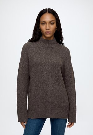 Femme aux longs cheveux foncés portant un pull marron en laine à col montant et un jean bleu, debout devant un fond clair uni.