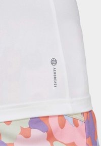 Bílý top z tkaniny AEROREADY bez vzorů, doplněný vícebarevnými, texturovanými šortkami s pastelovým kamuflážním vzorem.