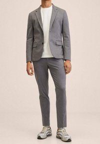 Man in een slim-fit grijs pak met een wit T-shirt en grijs-witte sneakers, staand tegen een beige achtergrond.