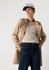 Gabardina beige con botones de doble botonadura, llevada sobre un suéter texturizado en color crema, combinada con jeans de pierna ancha en azul oscuro. Gorro blanco con logo verde.