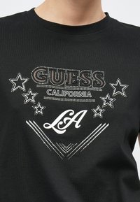 Zwart katoenen T-shirt met een voorkantgrafiek met "GUESS" in vetgedrukte letters, decoratieve sterren, strasstenen en "California LA" in cursief.