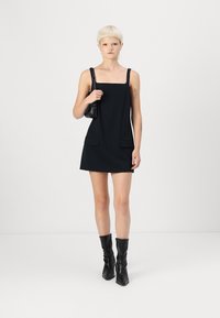 Robe noire sans manches avec un col carré et deux poches avant. Le modèle porte des bottes noires jusqu'au genou et tient un petit sac noir.