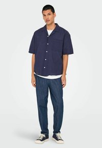 Cămașă navy cu mânecă scurtă, cu un buzunar, nasturi albi, croială lejeră, asortată cu pantaloni din denim blue și adidași închiși la culoare cu tălpi albe.