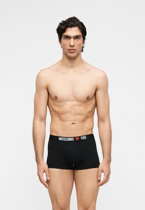 Homme torse nu aux cheveux foncés portant un boxer noir Moschino avec un logo blanc et rouge sur la ceinture, debout sur fond blanc.