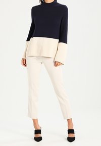 Navy- und crèmefarbener Farbblock-Pullover mit geripptem Muster und ausgestellten Ärmeln, kombiniert mit crèmefarbener Slim-Pants und schwarzen High Heels.
