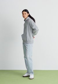 Grå huvtröja med framficka, i kombination med ljusblå jeans och vita sneakers, stående på en grön golv.