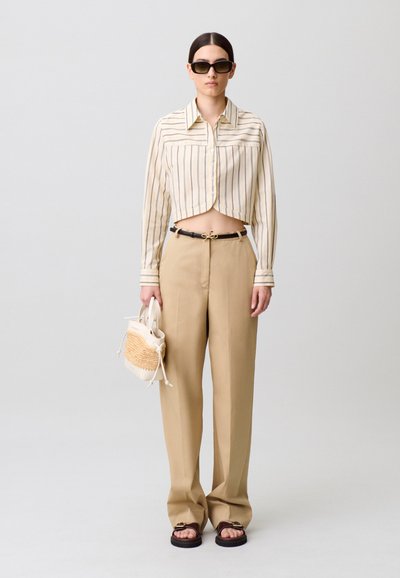 Strisce decorano una camicia cremisi corta con bottoni; abbinata a pantaloni beige a gamba larga e a una piccola borsa intrecciata. Il modello indossa occhiali da sole scuri.