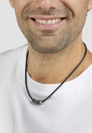 Schwarze Lederhalskette mit silbernen Metallakzenten, die drei dekorative Perlen aufweist und über einem weißen T-Shirt getragen wird. Glatte Struktur, lässiges Design.