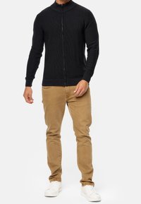 Mann in einem schwarzen Reißverschlusspullover, braunen Hosen und weißen Sneakers, der vor einem schlichten weißen Hintergrund steht.