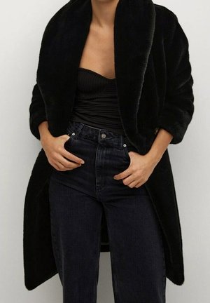 Personne portant un manteau en fausse fourrure noire par-dessus un top noir bustier et un jean noir taille haute, les mains reposant près des poches.