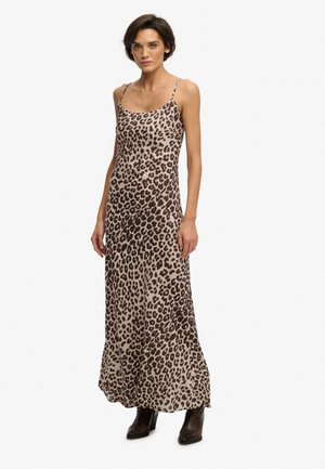Kvinde iført en ærmeløs, leopardmønstret maxikjole kombineret med brune ankelstøvler, stående mod en ensartet hvid baggrund.