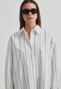 White button-up shirt with blue vertical stripes, featuring a classic collar and long sleeves. Accompanied by angular black sunglasses.