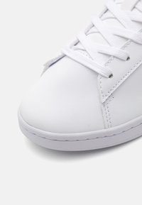 Hvit sneaker med glatt syntetisk overdel, rund tå og flat gummisåle. Har hvite snørebånd og subtile sømmer.