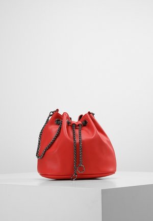 Czerwona skórzana torba typu bucket z sznurkiem do ściągania, czarnymi metalowymi łańcuszkami jako paskami oraz detalami w formie pierścieni, umieszczona na białym piedestale na jednolitym tle.