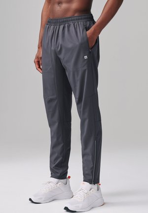 Homme portant un pantalon de sport gris foncé fuselé avec taille élastique et chaussures de course blanches, debout avec une main dans la poche.