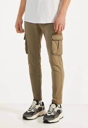 Homme portant un pantalon cargo beige avec de grandes poches latérales et des baskets épaisses noires et blanches, debout sur un fond blanc.