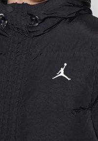 Zwarte pufferjack met een hoge hals, voorzien van een wit Jumpman-logo, een capuchon met trekkoord en een slanke, gestructureerde stof.