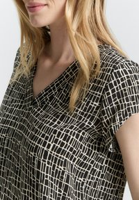 Schwarz-weiße Bluse mit kurzen Ärmeln und geometrischem Muster. V-Ausschnitt-Design aus weichem, fließendem Stoff mit leichter Textur.