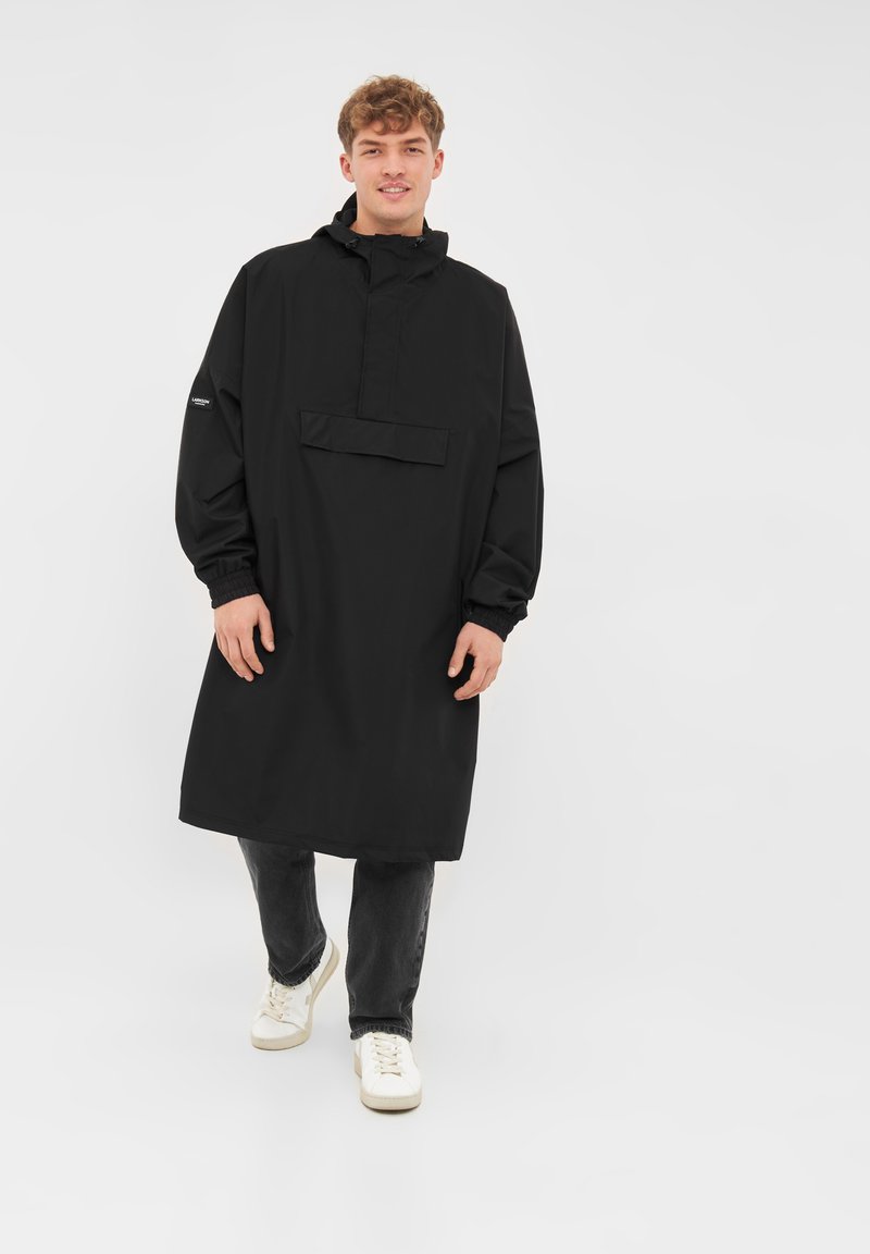 Jeune homme portant un long imperméable noir, un jean foncé et des baskets blanches, marchant vers l'avant sur un fond clair uni.