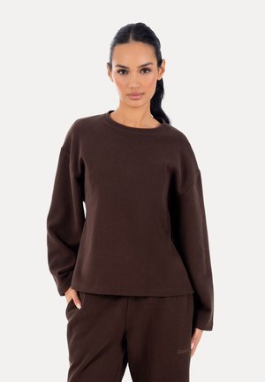 Kvinde med mørkt hår i en hestehale iført en løs mørkebrun langærmet top og matchende bukser, stående foran en ensfarvet lys baggrund.