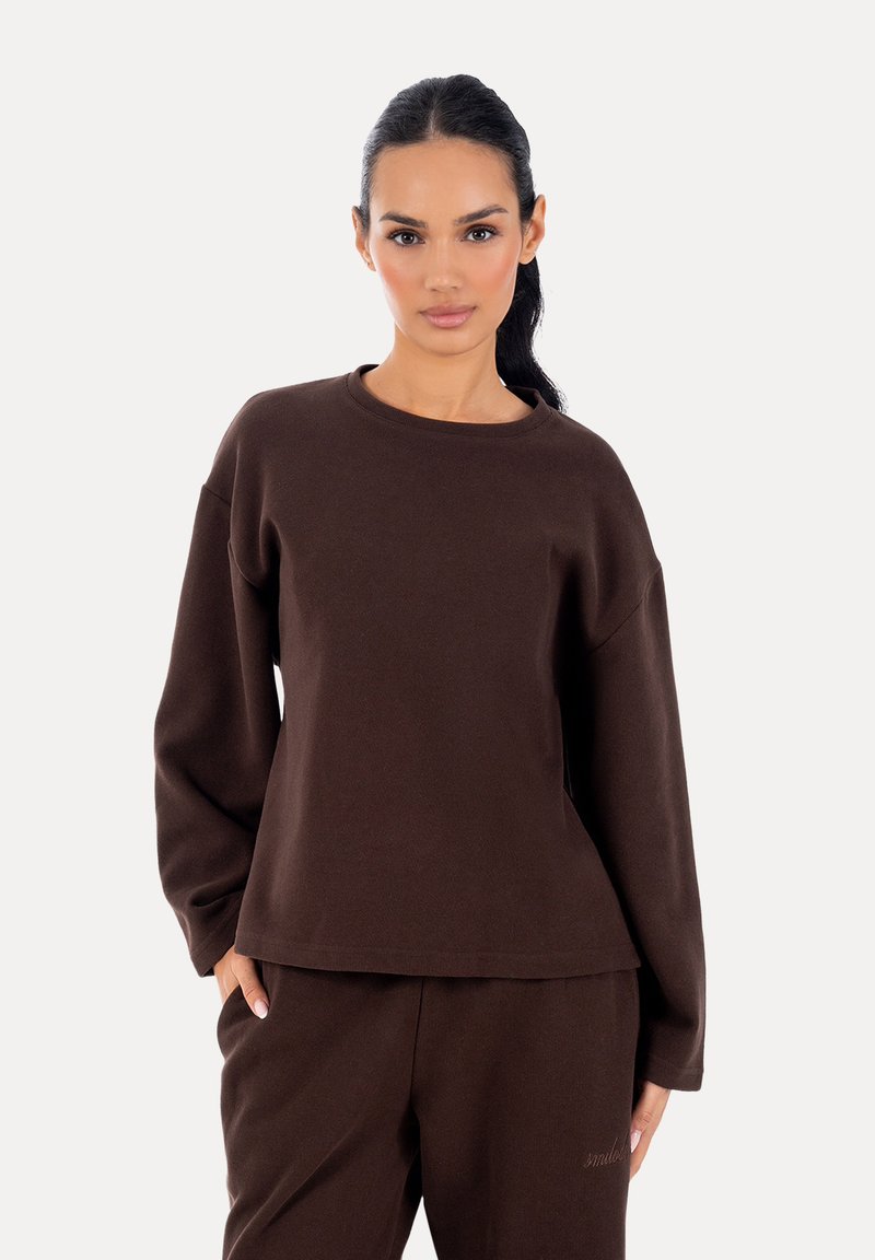Femme aux cheveux foncés attachés en queue de cheval, portant un haut ample à manches longues marron foncé et un pantalon assorti, debout devant un fond clair uni.
