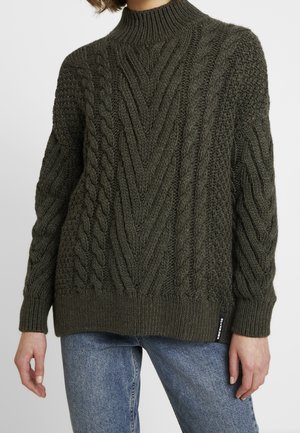 Frau trägt dunkelgrünen grob gestrickten Pullover mit hohem Kragen und blauen Jeans, steht vor einfarbigem Hintergrund.