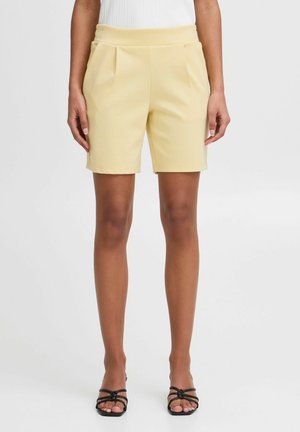Gule skræddersyede shorts med en glat tekstur, der har folder og en høj talje, parret med sorte strop sandaler.