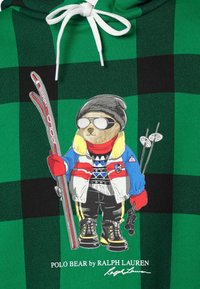 Zelená a černá kostkovaná mikina s kapucí, zdobená vyšitým medvědem v lyžařském vybavení, držícím lyže, se brýlemi, rukavicemi a barevnými akcenty.