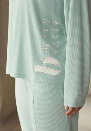 Close-up of person wearing light green textured long-sleeve top and pants with "BY TED BAKER" embroidered on the side.