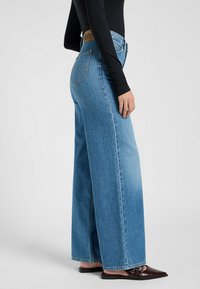 Høytlivte lyseblå vide jeans med en tan-skinnlogo og tradisjonelle baklommer. Brukt med en tettsittende svart langermet topp.
