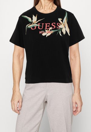 Sort t-shirt med kortærmer med "GUESS EST. 1981" og blomsterdesign, båret med lysegrå bukser af en stående person.