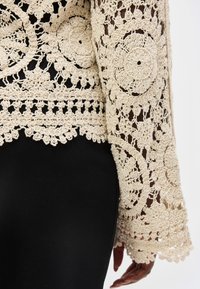 Rukáv krémovej háčkovanej čipkovanej odevnej látky s kruhovými kvetinovými vzormi na čiernom podklade a viditeľnou rukou po boku.