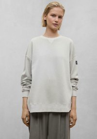 Sweatshirt gris clair avec des épaules tombantes, des poignets et un ourlet côtelés. Présente une petite étiquette noire sur la manche gauche. Tissu doux et texturé.