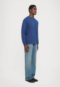 Sudadera azul marino con cuello redondo, corte relajado y detalle del logo en la parte frontal, combinada con jeans de pierna ancha de color azul claro y zapatos negros.