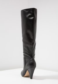 Schwarzes, knielanges Lederstiefel mit einem schlanken, geschwungenen hohen Absatz, der auf einer weißen Oberfläche vor einem schlichten hellen Hintergrund aufrecht steht.