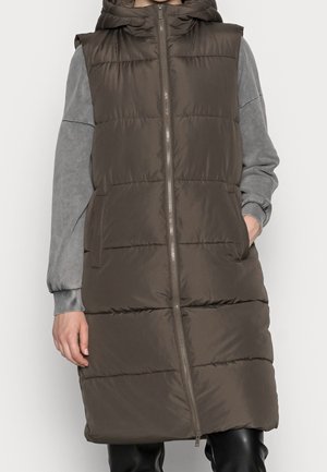 Personne portant une longue veste matelassée marron sans manches avec capuche sur un sweat-shirt gris, les mains dans les poches de la veste, debout devant un fond uni.
