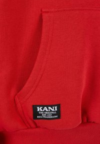 Červená mikina zo hrubej tkaniny s klokanou vreckom, pružnými manžetami a čiernou etiketou s bielym textom "KANI THE ORIGINAL EST. 1989."