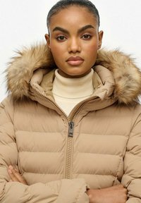 Chaqueta de plumas color beige claro con capucha con borde de piel, cierre de cremallera, diseño acolchado en forma de Q y cuello alto, llevada sobre un cuello de tortuga crema.