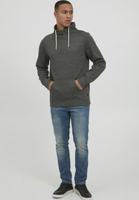 Grå hættejakke med høj krave, snoretræk og forlommer, parret med blå jeans og sorte sneakers på en ensfarvet baggrund.