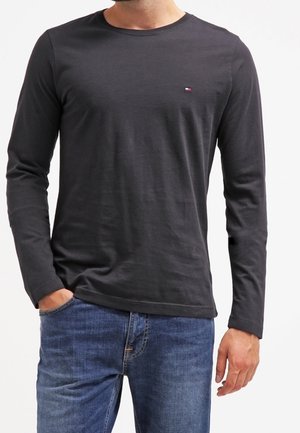 Mann trägt ein eng anliegendes schwarzes Langarmshirt mit einem kleinen Logo auf der Brust und blaue Jeans, eine Hand in der Tasche.